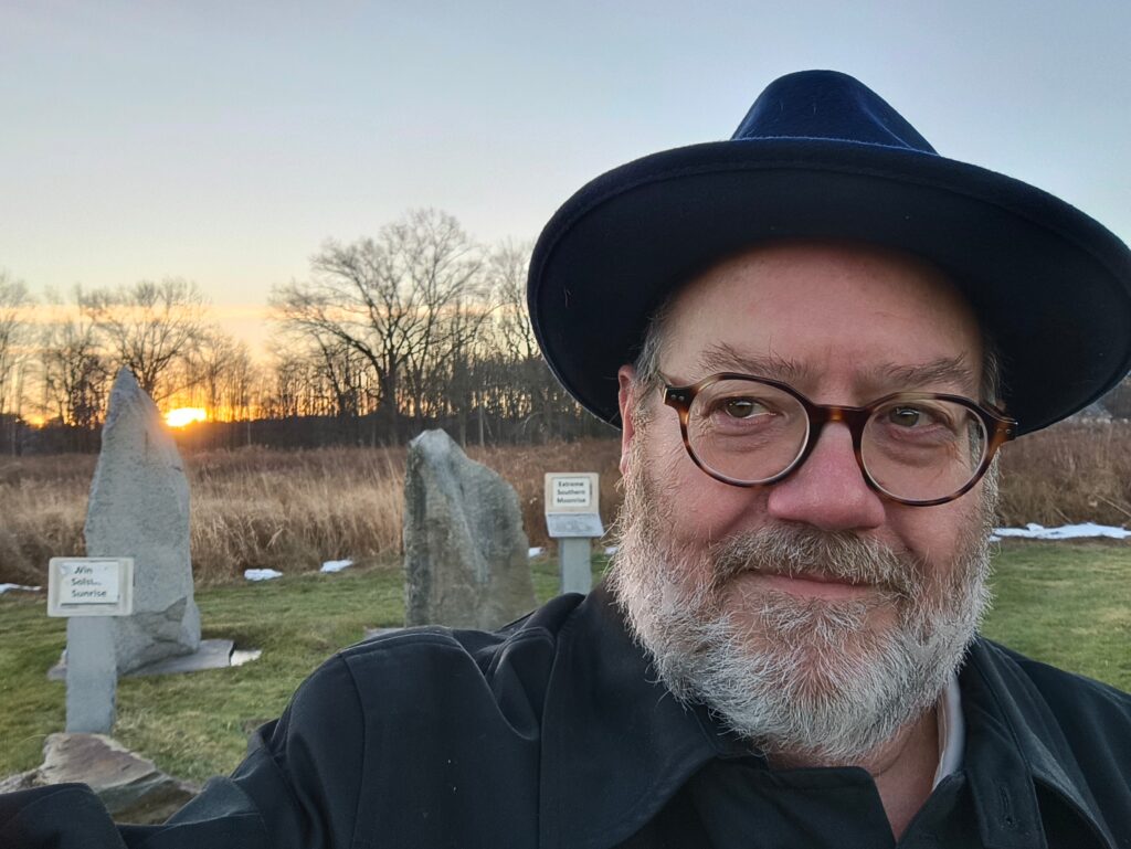 an older man wearing a hat in front of sun-aligned standing stones watching the sun rise on the winter solstice.