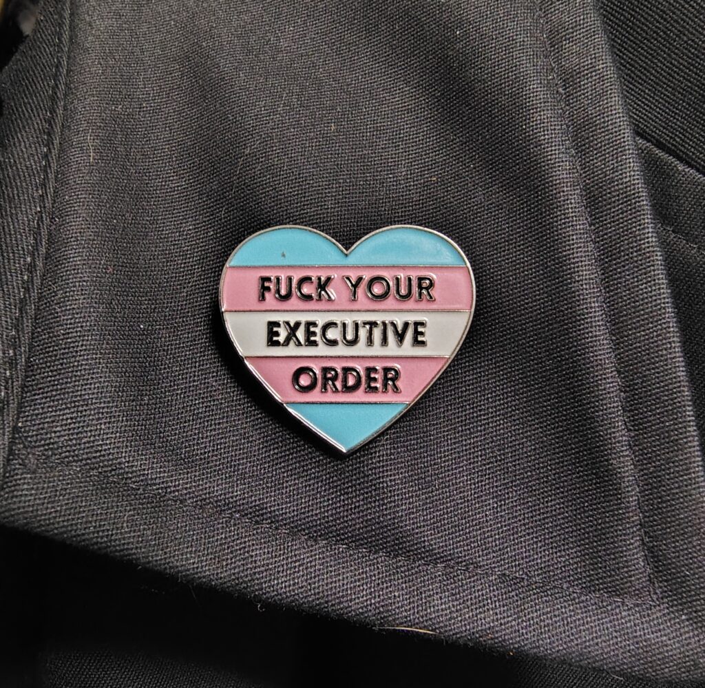 a heart-shaped pin with trans-flag colors that says, "FUCK YOUR EXECUTIVE ORDER."