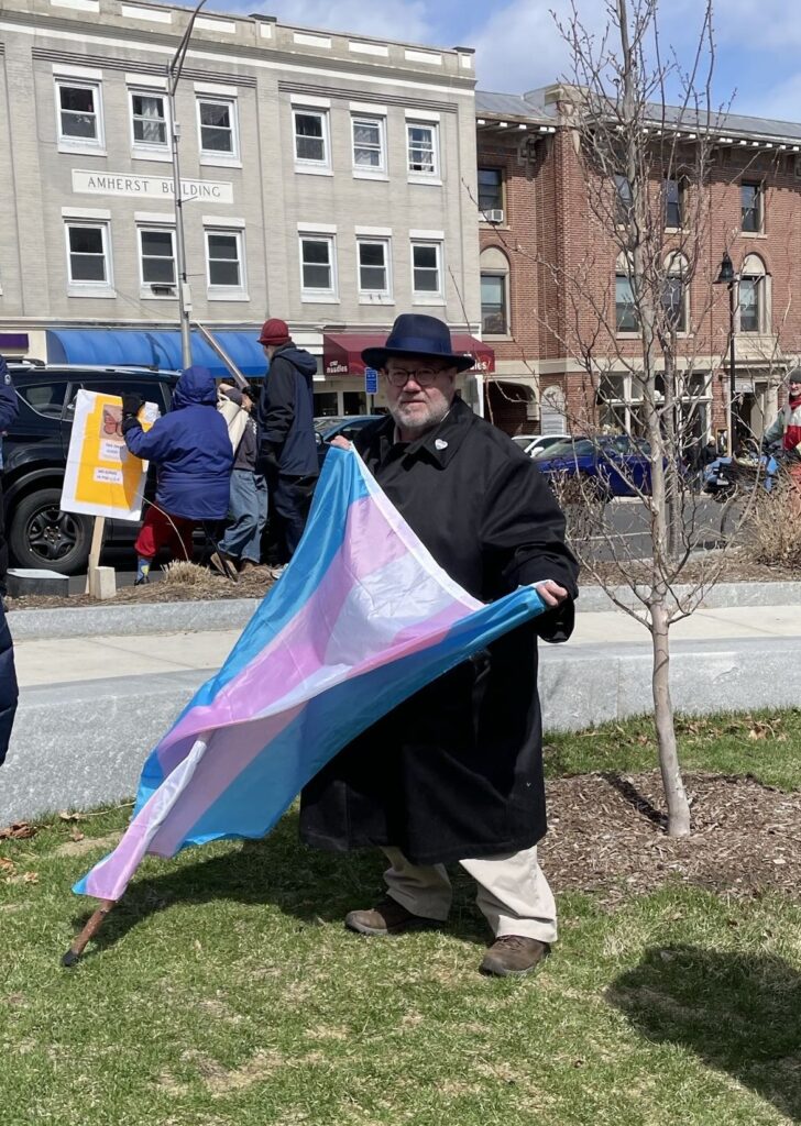 an older man wearing a trenchcoat and blue fedora is holding a trans flag at the Amherst No Kings protest.